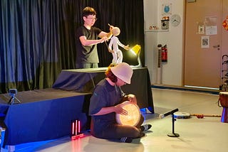 A puppeteer in black, moving a puppet in white using three black rods. A percussionist plays a lit drum while seated cross-legged on the floor.