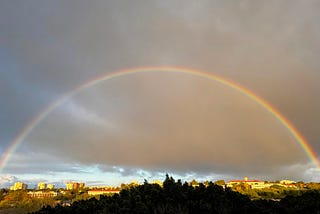 A full end to end rainbow with a dark-ish sky, and a few buildings padding the bottom edge