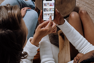 Two women scroll through a smartphone together from an overhead perspective.