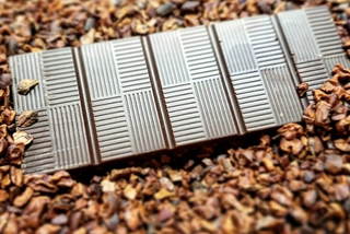 A chocolate bar, with five vertical segments with each segment with a pattern of shallow straight lines alternating vertically and horizontally in a quadrant layout, sitting on top of roasted cocoa nibs