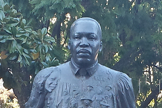 Dark colored head and shoulder statute of Martin Luther King, Jr. with trees in the background.