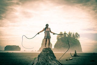A picture on a beach with the ocean behind and rocks rising from the shoreline. In the center foreground stands a brown-skinned woman on a rock on the beach. She is wearing a grey dress, sunglasses, and her hair is braided. She is holding kelp in each hand and wielding them like whips.