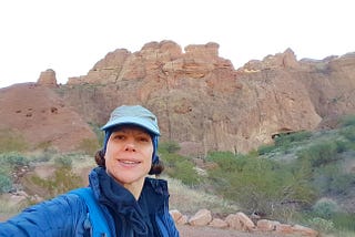 Woman wearing hat and head band covering her ears, high-neck shirt, jacket, with large rock in the distance