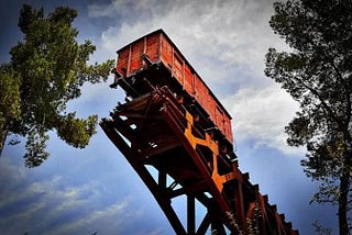 old orange train car suspended on broken track against blue sky with white clouds framed by leafy tree branches