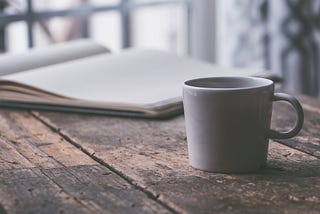 A cup of coffee and an opened book, all on a hardwood table.