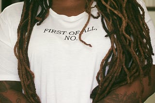 Close-up of defiant Black woman with her hands crossed in front of her wearing white tee with the words: FIRST OF ALL, NO.