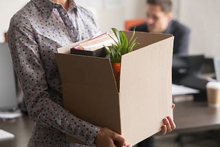 Person wearing a business shirt leaving an office, carrying a box filled with desk contents and a plant.