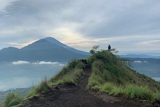 A black volcanic path surrounded by grass on a hill with a mountain in the background, light cloudy sky. a distant silhouet of a man stands at the edge of the cliff.