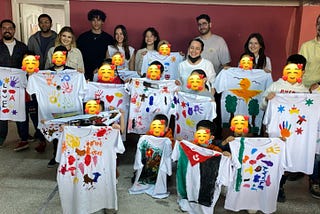 An photo of the Yasar student group with children holding the t-shirts they have painted. The children’s faces are covered with an emjoi.