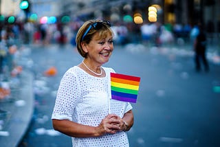 A white woman who looks to be young grandmother age, stands on a parade route, holding a small rainbow flag and smiling.