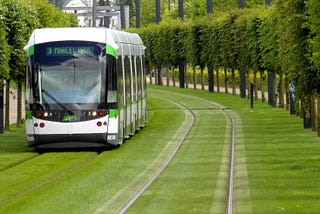 A tram travelling on a set of tracks, surrounded by grass and trees that form a beautiful green corridor through Nantes, France