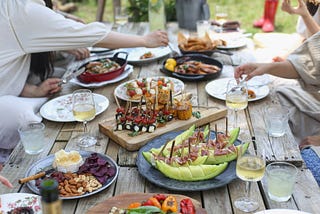 Wooden table on the grass covered with an array of delicious-looking food and drinks. Hands and arms reach across to help themselves. A pair of red boots appear on the grass in the corner.