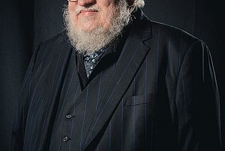 A photograph of George R.R. Martin in a suit. Attribution: Henry Söderlund, CC BY 4.0 <https://creativecommons.org/licenses/by/4.0>, via Wikimedia Commons