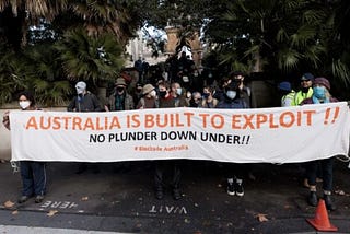 Crowd of people holding banner reading Australia is built to exploit!! No plunder down under #BlockadeAustralia