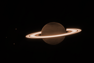 Dark brown Saturn surrounded by bright glowing rings. Three planets sit on the left hand side of it.
