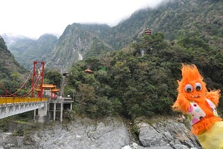 An Orange puppet with googly eyes and a polka-dot vest sticks out over Taiwan’s Taroko Gorge. Behind him an orange and red bridge spans the chasm. Behind this, a pagoda and the Chinese goddess Kuanyin sit among the mountains.