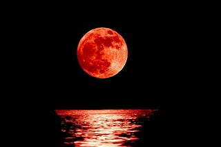 A picture of a blood red moon rising above the water.