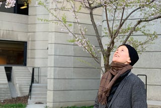 Photo of the poet and writer looking up into the cherry blossom trees. They are wearing a grey jacket and a black hat.