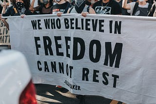 Protesters carrying large white sign with black text “We who believe in Freedom cannon rest”