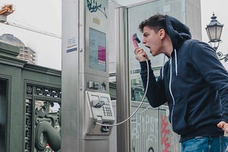 This is an image of a white guy in a jeans and a dark blue hoodie who is screaming into the receiver of a payphone and gesturing angrily.