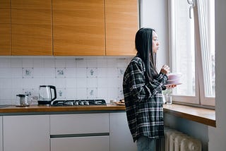 A dark-haired woman stands alone in her kitchen, holding a bowl, and looking out the window. A cooktop and coffee pot are visible behind her, and a radiator is under the windowsill through which she looks at the world.