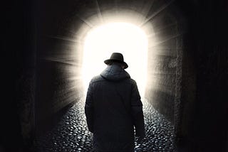 Silhouette of the back of a man in a heavy coat and fedora walking through an arched doorway from darkness into the light. Depositphotos | frankie_s