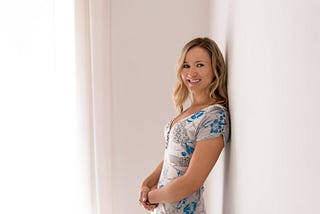 Blonde woman in blue, grey and white dress leaning against white wall