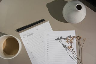 Note pages pre-formatted for checklists of different lengths. Remnants of a light coffee to the left, dried flowers and a vase to the right. The pen rests, capped, across the top of the pages.