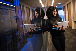 The image shows a woman leaning against a glass wall separating her work area from a room full of servers and other computer equipment. The woman has a laptop on her lap and appears to be focused on her work. Behind her, servers and network equipment are visible. The image evokes the feeling of working in a modern, tech-savvy space surrounded by state-of-the-art computer devices.