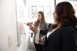 Two women standing at a whiteboard writing on it and working together