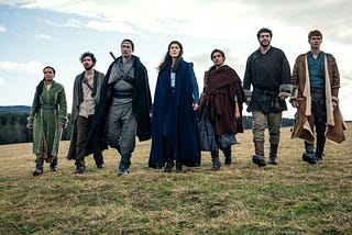 Nynaeve, Mat, Lan, Moiraine, Egwene, Perrin, and Rand walk together, side-by-side, across a field.