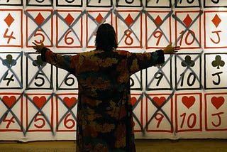 A man wearing a robe stands, arms out, in front of a wall with playing cards painted on. Most of them have a black X painted.