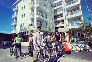 Picture of Little Mountain Cohousing building from the alley. Five people pose on bicycles, in front of a fence. Behind that is a six-story, L-shaped building around a courtyard.