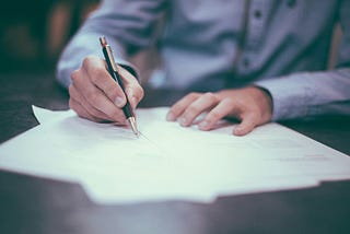 A man’s hand holds a pen. He is signing some documents that are on a table.