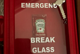 Picture of gag gift featuring a hammer and a packet of ketchup behind glass with “emergency break glass” written on it.