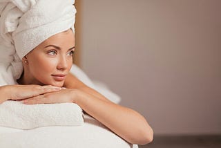 Image shows a woman lying on a massage table. Her hair is pulled up into a towel on top of her head and she is lying on her stomach, arms supporting her chin. Her eyes are open and she looks relaxed.
