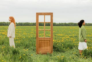 Two women standing away from each other in a field, with a free standing door between them.