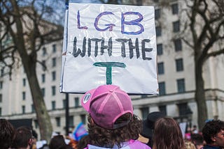 Building in the background. Protest sign held by person with brown curly shoulder length hair, wearing a pink snap back. The Sign is a white rectangle that says ‘L G B with the T’.