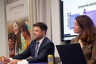 World Learning’s Vice President for External Engagement and Advocacy Joel Colony sits at a table next to a woman. Behind them is a World Learning banner and a projection of a presentation.