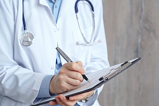 A doctor wearing a white lab coat and stethoscope holds a clipboard and pen.