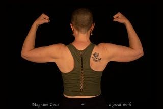 A photograph taken in a studio with a black background, a woman with a shaved head facing backwards with both arms flexed showing her muscles; she has a black and white tattoo on her right shoulder that is an angel with two words written below it: “Magnum Opus.” There is text in white at the bottom that reads “Magnum Opus = a great work.”