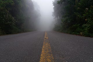 Foto de estrada asfaltada deserta, cercada de árvores. A linha amarela desgastada segue até o horizonte, acabando em neblina