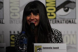 Jameela sits in front of a black and white background, smiling into mic, wearing black and white blouse