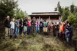 A group of people dressed for outdoor work stand in a semi-circle with linked arms, smiling, with a big log home behind them