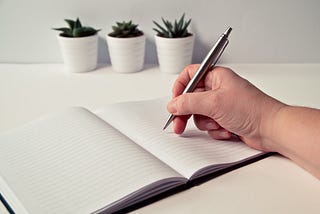 A photo of a hand holding a pen and writing in a blank book.