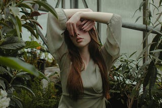 A woman in a soft green dress stands in a greenhouse surrounded by foliage, covering part of her face with her hands. Her expression is obscured, suggesting introspection or emotional overwhelm.