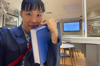 印刷された博士論文を抱える筆者の自撮り。A4サイズ400ページ、青のハードカバーの論文を左手で抱えて右手を伸ばして自撮りした。