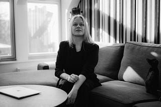 Portrait of Executive Life Coach Signe Jungløw in black and white sitting in a sofa bathed in sunlight with a black kitten on her side and a macbook air on the table in front of her. She is wearing all black.
