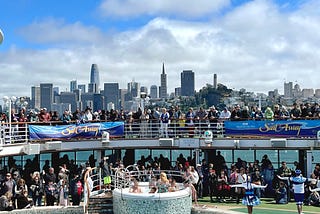 Image of a crowded group of guests and crew dancing and celebrating on the pool deck (level 15) and upper deck (level 16) of the Ruby Princess cruise ship with the San Francisco skyline in the background below a bright blue sky and fluffy white clouds. Two large banners hanging from the guardrails read “Sail Away” party.