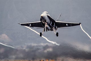 A photograph of a jet fighter with its wheels still down after having just lifted off from the runway. Vapour trails are being created by its wingtips.